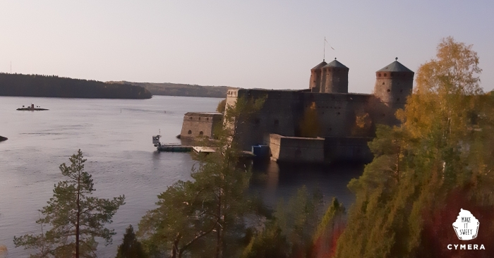 Kuvassa on näkymä Olavinlinnaan linnan virran toisella puolella olevan kallion päältä, josta linna näkyy Saimaa taustalla.